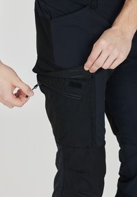 Svarta multifunktionella byxor med blixtlåsfickor, tillverkade av ett slitstarkt, texturerat tyg, med en kombination av paneler och justerbara stängningar.