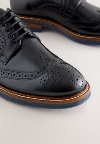 Black leather dress shoes with perforated wingtip design, lace-up closure, and a contrasting brown welt on a blue rubber sole.