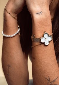 Une montre avec un bracelet en maillon doré présente un cadran en forme de trèfle à quatre feuilles blanc avec des accents dorés. Un bracelet en perles orne le poignet aux côtés de tatouages.