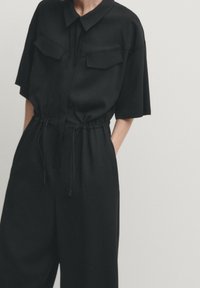 Zwart kortemouwen jumpsuit met een kraag, twee borstzakken, een elastische taille met trekkoorden en wijde benen. Zachte textuur.