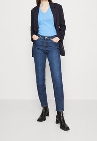 Džíny z denimu v tmavě modré barvě, spárované se světle modrým topem s výstřihem do V a černým sakem. Modelka stojí s rukama v kapsách, na sobě má černé boty na podpatku.