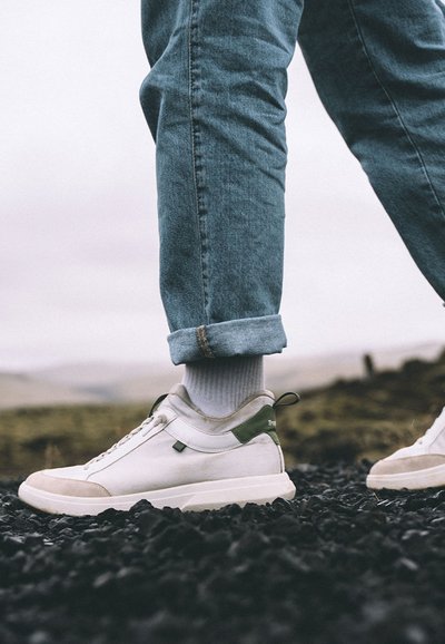 Baskets blanches avec des accents verts, embout en gris clair et dessus en textile. Assorties à un pantalon en denim retroussé et des chaussettes blanches. Le sol est sombre.