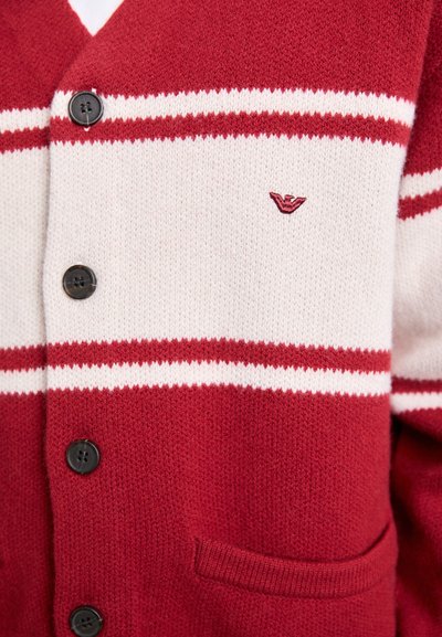 Cardigan tricotat cu dungi roșii și albe, cu butoane negre, un mic logo brodat și o buzunar frontal.