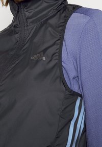 Svart ärmlös jacka med hög krage, tillverkad av glänsande material, har en diskret Adidas-logotyp och ljusblå sidostrepen.