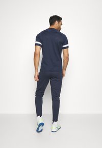 Marineblå atletikoutfit bestående af en kortærmet top med hvide striber og tætsiddende bukser, lavet af åndbart materiale, med lynlåse ved anklerne.
