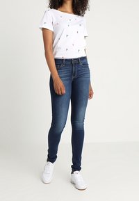 Camiseta blanca con detalles bordados coloridos, combinada con jeans ajustados de color azul oscuro y zapatillas blancas. Diseño suave y ceñido en general.