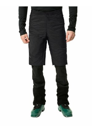 Shorts noirs isolés avec une surface texturée, associés à un pantalon noir ajusté. Des chaussures de ski vertes complètent la tenue.