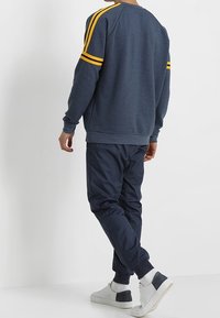 Felpa navy con strisce gialle sulle spalle, abbinata a pantaloni jogger navy scuri e sneaker bianche, caratterizzata da una texture liscia e una vestibilità casual.