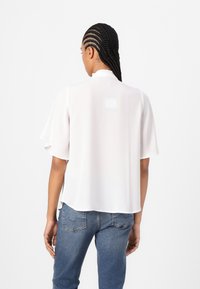 Blusa bianca a maniche corte con una leggera lucentezza, colletto arricciato e design morbido, abbinata a jeans blu.