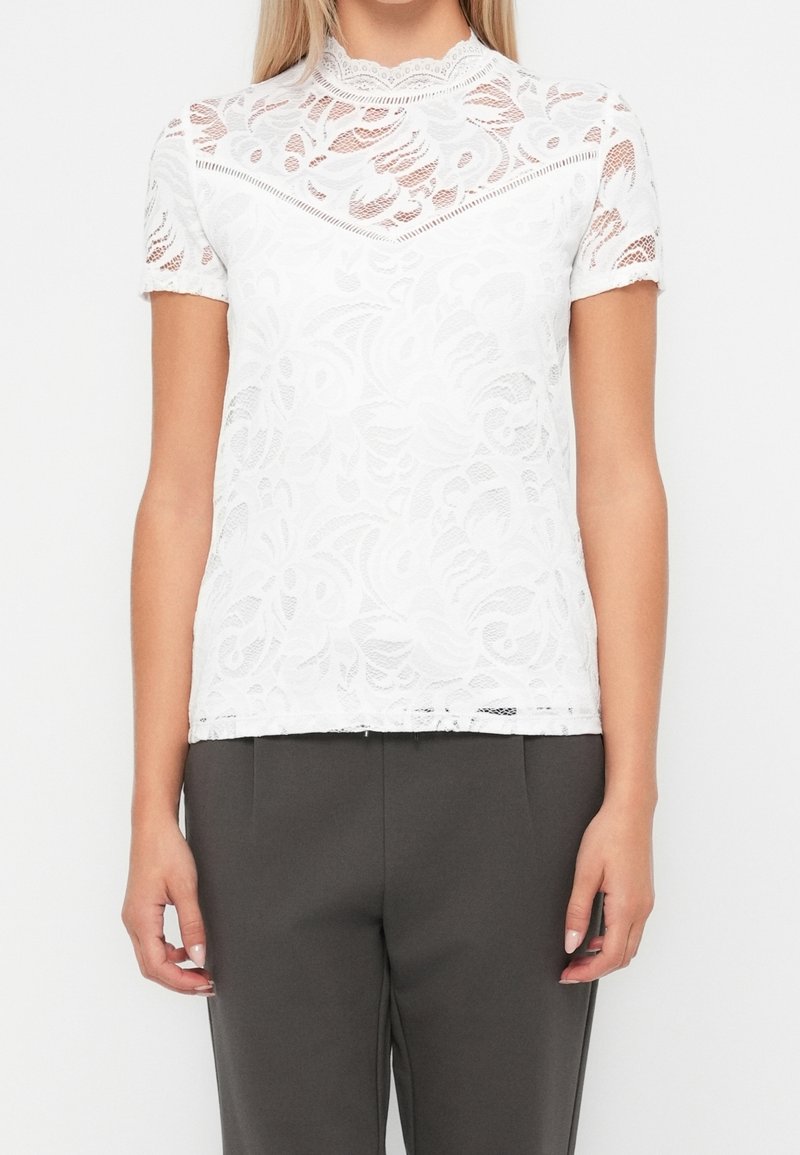 Femme portant un chemisier blanc en dentelle à manches courtes avec des motifs floraux et un col montant, associé à un pantalon gris foncé.