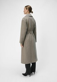 Abrigo largo con cinturón, hecho de tejido gris texturizado, con un cuello tipo solapa y cortes laterales, combinado con jeans negros y tacones afilados.