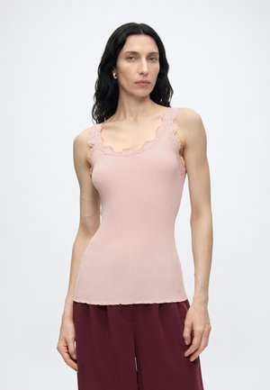 Femme aux cheveux noirs ondulés portant un haut sans manches rose pâle avec bordure en dentelle et un pantalon large rouge foncé, debout devant un fond uni.