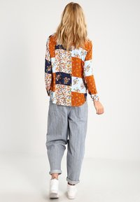 Langarm patchwork shirt met bloemen- en geometrische patronen in oranje, blauw en wit, gecombineerd met lichtgestreepte, baggy broeken.