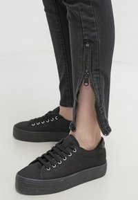 Baskets noires associées à un jean skinny noir avec des fermetures éclair ouvertes aux chevilles révélant la peau sur un fond clair.