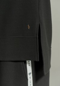 Czarny top z teksturowanej tkaniny, z prążkowanym wzorem, zamkowymi bokami i małym złotym akcentem w postaci logo. Zawiera widoczny metek na dolnym brzegu.