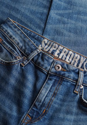 Denim jeans i en mellem blå vask, med kontraststing, en mærket talje med "SUPERDRY" og hardware i kobberfarve.