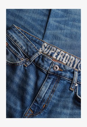 Jean de denim en un lavado azul medio, con costuras en contraste, una cinturilla con la marca "SUPERDRY" y herrajes en tono cobre.