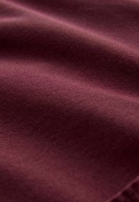 Tejido burdeos con una textura lisa y ligeramente brillante. La superficie presenta una sutil veta y un color uniforme, creando una apariencia suave y fluida.