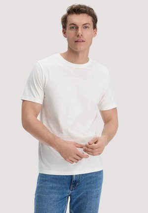 Giovane uomo con una semplice maglietta bianca a maniche corte e jeans blu, in piedi davanti a uno sfondo neutro, guarda avanti con le mani vicino alla vita.