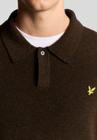 Σκούρο καφέ πλεκτό πόλο με γιακά με κουμπιά και κίτρινο κεντημένο λογότυπο στο αριστερό στήθος. Υφασμάτινη υφή, φορεμένο με ένα λευκό φανελάκι από μέσα.