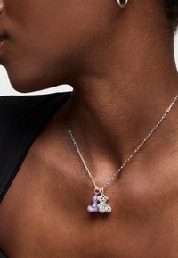 Collier en chaine en argent avec trois breloques : une perle violette, une perle en filigrane argentée, et une forme en perles, reposant sur un haut sombre.