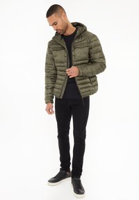 Chaqueta impermeable verde oliva con capucha, que presenta un cierre de cremallera y dos bolsillos frontales. Lleva puesta sobre una camiseta negra y combinada con jeans negros y zapatillas.