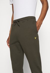 Olivgrüne Sweatpants aus weichem Stoff, mit einem elastischen Bund und einer Kordel, sowie einem kleinen gelben gestickten Logo an der Seite.