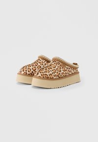 Paire de chaussures sans lacets avec tige imprimée léopard beige, semelles compensées épaisses beige, et logo UGG sur le côté, présentées sur fond blanc.