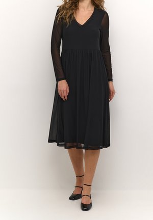 Kobieta ubrana w czarną sukienkę midi z przezroczystymi długimi rękawami i dekoltem w serek, w połączeniu z czarnymi szpilkami na pasku wokół kostki, stojąca na jednolitym tle.