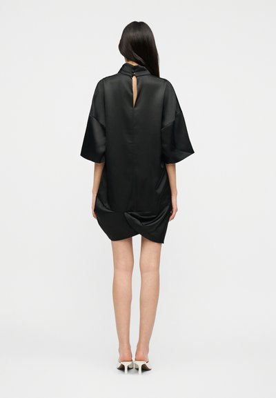 Γυναίκα με μακριά σκούρα μαλλιά που φοράει ένα χαλαρό μαύρο σατέν μίνι φόρεμα με φαρδιά μανίκια και λευκά παπούτσια με ψηλό τακούνι, στέκεται με την πλάτη στραμμένη.