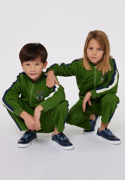 Tute da ginnastica verdi con accenti blu scuro, giacche con zip e design di elefante ricamato. Ogni bambino indossa scarpe slip-on blu scuro con elementi decorativi.