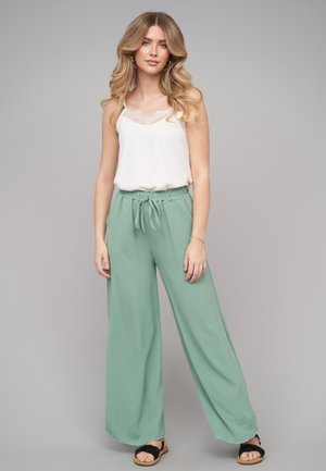 Junge Frau mit langen, welligen Haaren, die ein weißes Spitzen-Top, lockere mintgrüne Hosen und schwarze Sandalen vor einem grauen Hintergrund trägt.