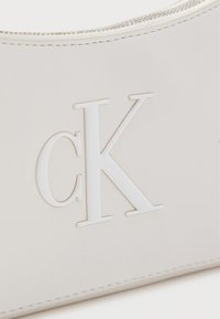 Λευκό δερμάτινο αντικείμενο με ανάγλυφο λευκό λογότυπο "cK" και ορατό μεταλλικό φερμουάρ κατά μήκος της πάνω άκρης.
