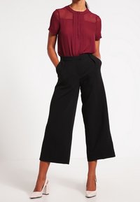 Blouse courte maroon à manches courtes avec des détails de boutons et un tissu transparent, associée à un pantalon noir à jambes larges et des chaussures à talons carrés de couleur claire.