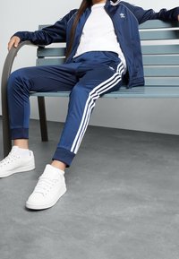 Navy tracksuit with white stripes, featuring a zip-up jacket and tapered pants, paired with white sneakers, seated on a bench.