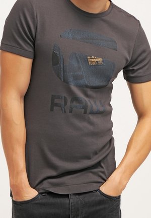 Mann trägt ein dunkelgraues T-Shirt mit einem großen, runden Jeansflicken und dem aufgedruckten Schriftzug "RAW" auf der Vorderseite, Hände in den Hosentaschen aus Jeans.