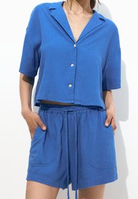 Camisa de botones de manga corta azul con cuello, combinada con pantalones cortos de cordón a juego que cuentan con grandes bolsillos laterales y un ajuste relajado.