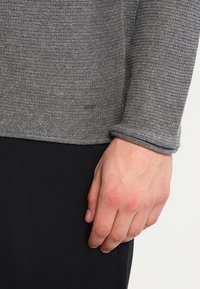 Pull gris en tricot avec une finition texturée et des poignets retroussés, comportant une étiquette visible à l'ourlet inférieur. La main du mannequin repose sur le côté.