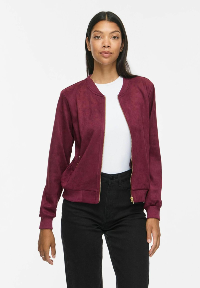 Femme portant une chemise blanche, un pantalon noir et une veste bomber bordeaux ouverte, debout devant un fond blanc.