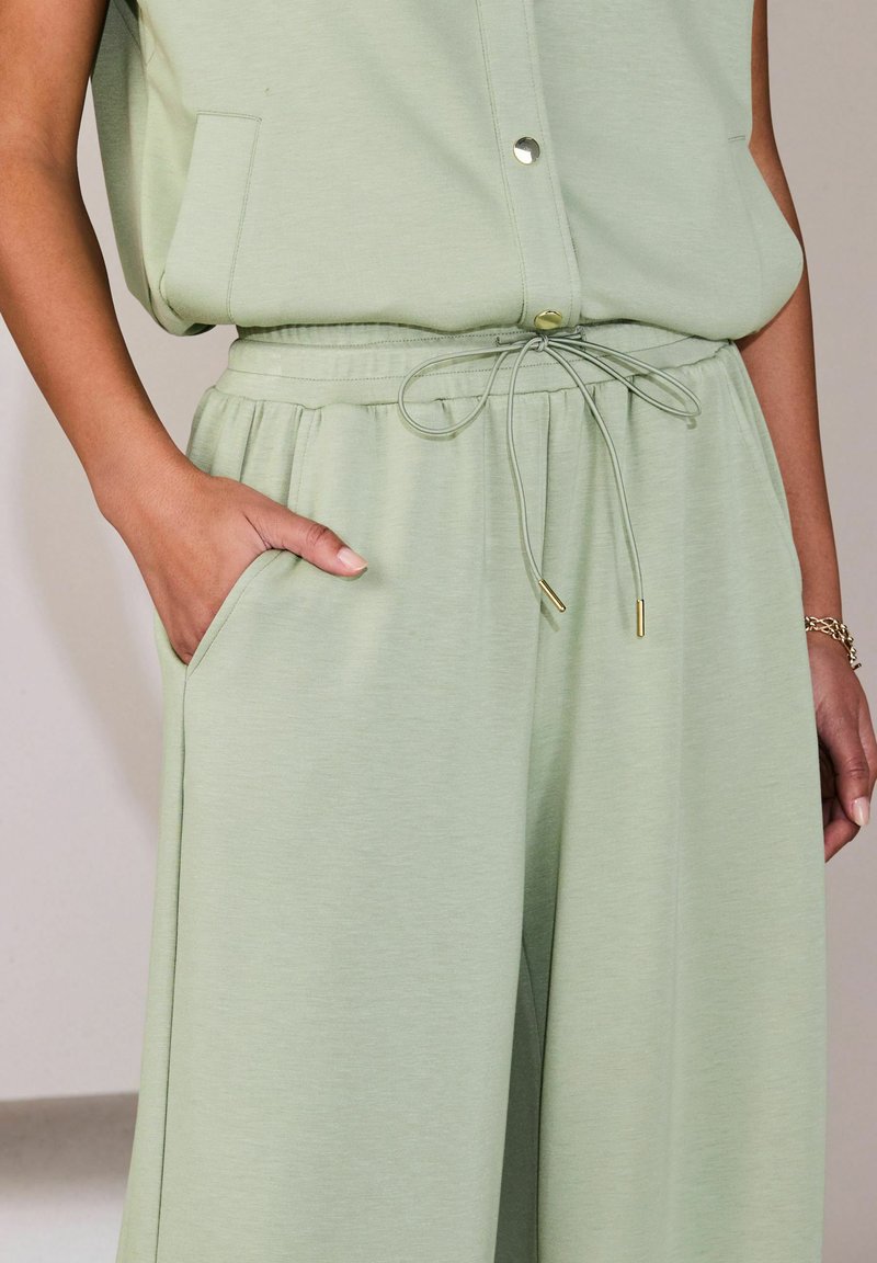 Femme portant un débardeur à boutons vert clair et un pantalon à cordon de serrage assorti, avec une main dans la poche et un bracelet au poignet.