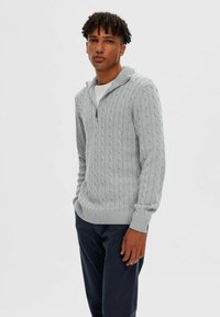 Hellgrauer, grob gestrickter Pullover mit halbem Reißverschluss am Kragen sowie gerippten Bündchen und Saum, getragen über einem weißen T-Shirt und kombiniert mit dunklen Hosen.