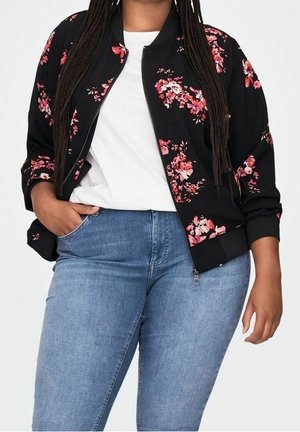 Persoon draagt een zwarte jas met bloemenprint en rits over een wit shirt, gecombineerd met lichtblauwe spijkerbroek, met hand in de zak, staand tegen een effen achtergrond.