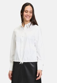 Camicia bianca con colletto e dettagli in argento, orlo arricciato con coulisse e maniche lunghe. Indossata sopra una gonna di pelle nera.