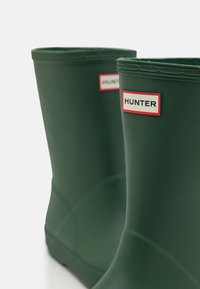 Stivali di gomma verdi con finitura opaca, dotati di un logo "HUNTER" rosso e bianco. Il design è dritto con un bordo superiore arrotondato.