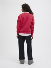 Sweatshirt rouge à coupe décontractée, ourlet arrondi et poignets côtelés, porté par-dessus une chemise rayée. Associé à un denim large de couleur foncée et des chaussures retroussées.
