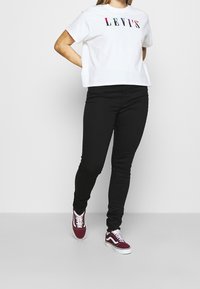 Camiseta blanca corta con el logotipo "LEVI'S" multicolor, combinada con jeans negros ajustados y zapatillas burdeos con acentos blancos.