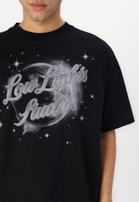 Must musta T-särk, millel on graafiline disain planeedist ja tähtedest, koos tekstiga "Low Lights Studios" hõbe- ja valgetes toonides.