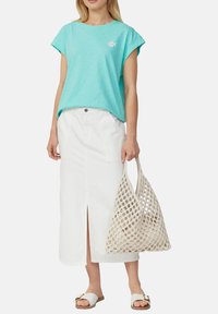 Vrouw in een turquoise t-shirt met korte mouwen, een witte midirok met een split aan de voorkant, witte sandalen en een beige netten tote bag.