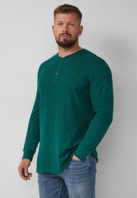 Türkisfarbendes Langarm-Henley-Shirt mit drei Knöpfen, strukturierter Oberfläche und aufgesetzter Tasche am Oberarm. Getragen mit hellblauen Jeans.