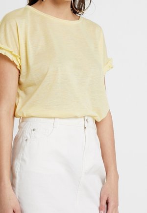 Kvinde iført en lys gul kortærmet top med op-rullede manchetter, stoppet ned i højttaljede hvide bukser mod en hvid baggrund.